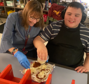 A teacher and student with headphones sit at a table and smile while putting a cheese topping on mini pizzas.